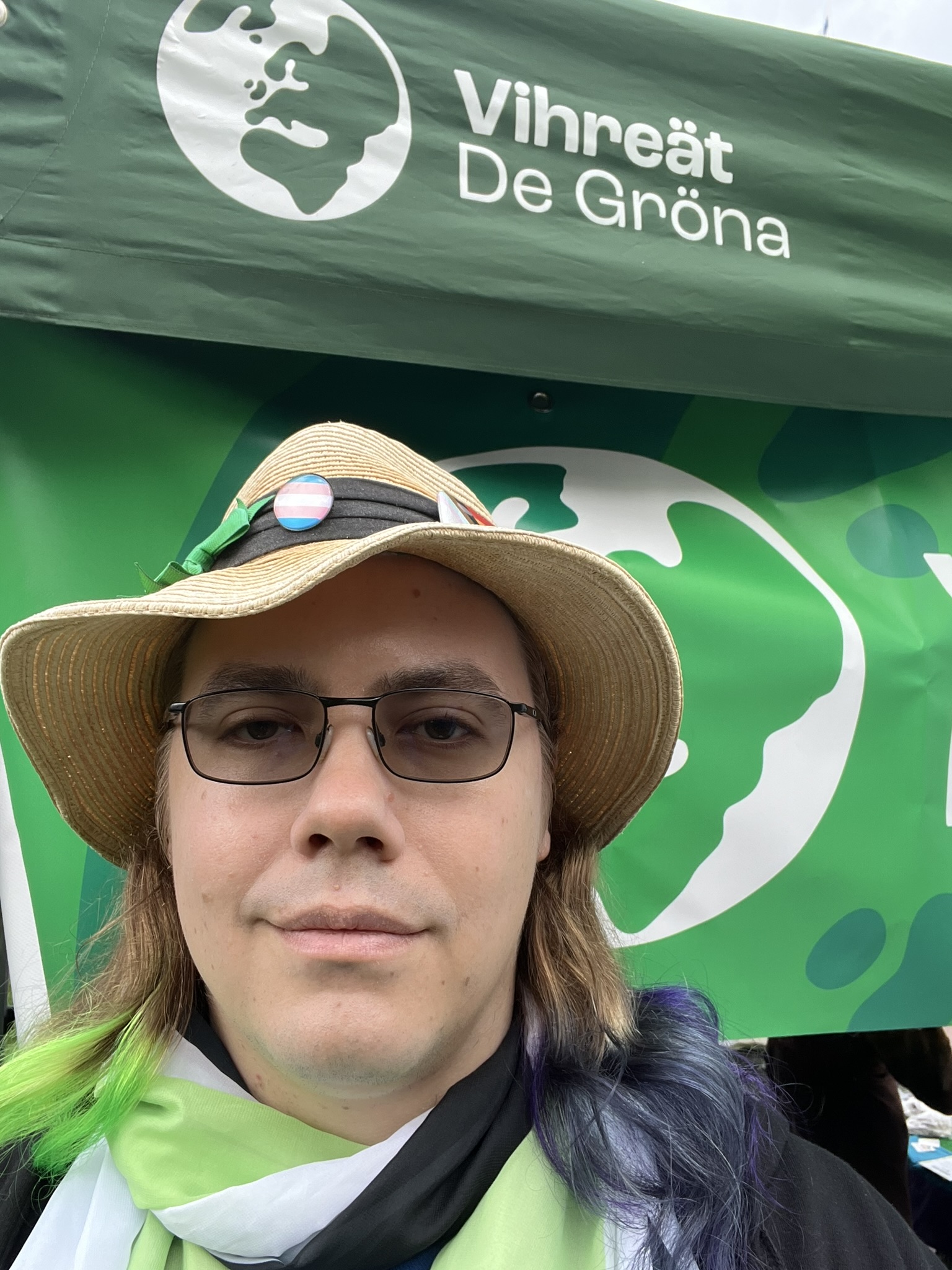 Selfie of me in front of the Green Party tent. On my hat is a trans flag pin; I am also wearing a scarf with the agender flag and my hair is dyed (visibly) black, purple, green and a bit of yellow, i.e. the nonbinary flag plus green.